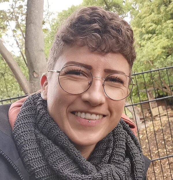 Selfie einer Absolventin der Euro Akademie Berlin. Sie trägt eine große modische und gemusterte Brille, braune kurze Haare, mit sehr kurzen Seiten, sie lächelt sehr freundlich, trägt einen grauen Strickschal, im Hintergrund sieht man ein waldähnliches Gelände.