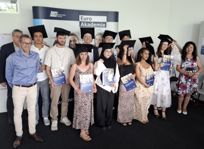 Absolventinnen und Absolventen der Euro Akademie Mainz posieren, alle mit eckigem Absolventen-Hut, und Lehrkräften gemeinsam für ein Gruppenfoto.