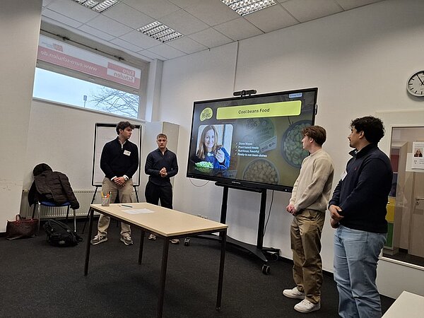 Vier junge Männer stehen in einem hellen Seminarraum vor einem großen Bildschirm. Der Bildschirm zeigt eine Präsentationsfolie mit dem Titel 'Coolbeans Food' und einem Porträt einer Frau. Auf der Folie sind außerdem einige kurze Stichpunkte und ein Bild von Lebensmitteln zu sehen. Die Männer tragen Freizeitkleidung und stehen in einer Reihe, während sie sich auf die Präsentation konzentrieren. Im Hintergrund sind ein Fenster, eine Uhr und eine weiße Wandtafel sichtbar. Die Atmosphäre wirkt sachlich und konzentriert.