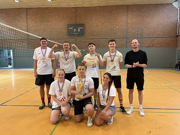 In einer Sporthalle stehen acht Personen auf einem orangefarbenen Hallenboden vor einem Volleyballnetz. Sie tragen Medaillen um den Hals und sportliche Kleidung, überwiegend weiße T-Shirts. Zwei Personen knien vorne, halten einen goldenen Pokal. Die übrigen stehen dahinter, einige zeigen Siegergesten. Im Hintergrund sind eine Anzeigetafel mit Zahlen und eine Backsteinwand zu sehen.