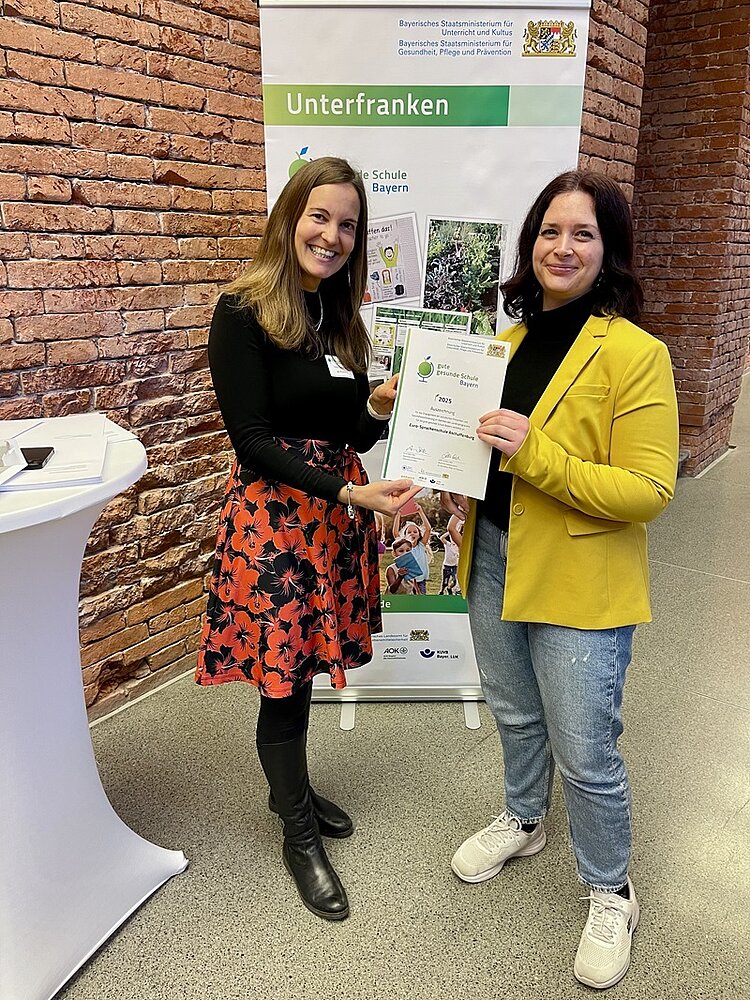 Die pädagogische Leitung der Euro Akademie Aschaffenburg, Vanessa Abele, nimmt in München die Auszeichnung zur „Guten gesunden Schule Bayern“ entgegen. Zwei Frauen stehen vor einem Banner des Bayerischen Staatsministeriums und halten gemeinsam und lächelnd die Urkunde in den Händen und in die Kamera für ein Foto. 