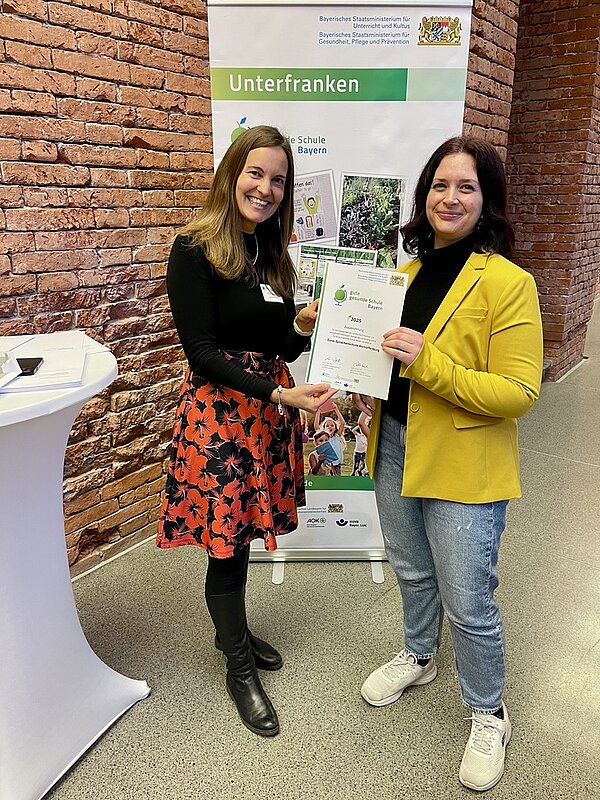 Die pädagogische Leitung der Euro Akademie Aschaffenburg, Vanessa Abele, nimmt in München die Auszeichnung zur „Guten gesunden Schule Bayern“ entgegen. Zwei Frauen stehen vor einem Banner des Bayerischen Staatsministeriums und halten gemeinsam und lächelnd die Urkunde in den Händen und in die Kamera für ein Foto. 