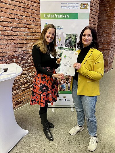 Die pädagogische Leitung der Euro Akademie Aschaffenburg, Vanessa Abele, nimmt in München die Auszeichnung zur „Guten gesunden Schule Bayern“ entgegen. Zwei Frauen stehen vor einem Banner des Bayerischen Staatsministeriums und halten gemeinsam und lächelnd die Urkunde in den Händen und in die Kamera für ein Foto. 