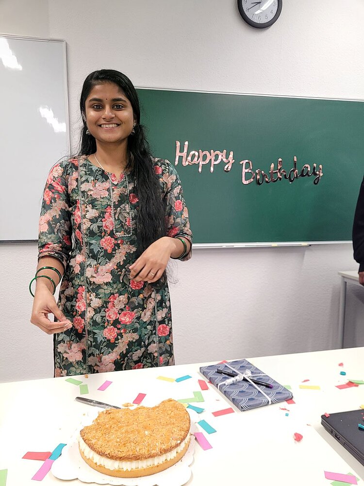Erfolgreiche Integration unserer indischen Auszubildenden Eine junge Frau mit langen schwarzen Haaren und einem floralen Kleid steht lächelnd vor einer grünen Tafel, auf der „Happy Birthday“ in glänzenden Buchstaben geschrieben steht. Sie scheint dabei zu sein, ein Stück Kuchen zu schneiden. Auf dem Tisch vor ihr liegt ein halb geschnittene Torte mit Sahnefüllung, umgeben von bunten Konfettistreifen. Daneben befindet sich ein verpacktes Geschenk mit einer Schleife sowie einem Laptop. Im Hintergrund sind ein Whiteboard und eine Uhr an der Wand zu sehen.