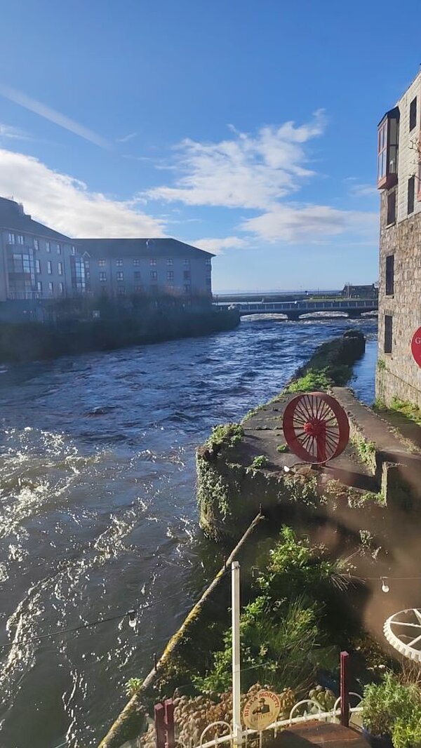 Ein Fluss oder Meerarm fließt durch eine Stadt mit historischen Gebäuden und einer alten Mühle oder einem Wasserrad.