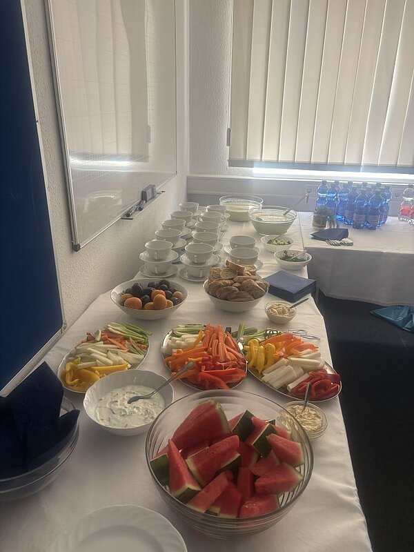 Ein langes Buffet mit einer Vielzahl von Snacks und Getränken. Dazu gehören Obst wie Aprikosen, Trauben und Wassermelonenstücke, Gemüsesticks mit Dips, Kekse, Muffins und Flaschen mit Wasser. Alles ist ordentlich auf einem Tisch mit weißer Tischdecke arrangiert.
