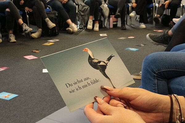 Theorie meets Praxis Eine Person hält eine Karte mit dem Bild eines Vogels, der an einen Sekretärvogel erinnert, und dem Text "Du siehst aus, wie ich mich fühle." Die Person sitzt in einem Stuhlkreis mit mehreren anderen Menschen, die im Hintergrund zu sehen sind. Auf dem Boden liegen verschiedene Karten verteilt.