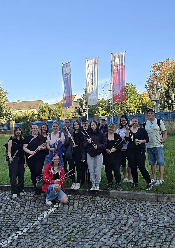 Die Schülerinnen und Schüler der Euro Akademie Hohenstein-Ernstthal posieren gut gelaunt draußen auf dem Parkplatz vor der Schule für ein Gruppenfoto. Sie halten alle eine Müllzange in den Händen. Im Hintergrund wehen große Fahnen der Euro Akademie und der Euro-Schulen an den Masten. Der Himmel ist wolkenlos blau. 