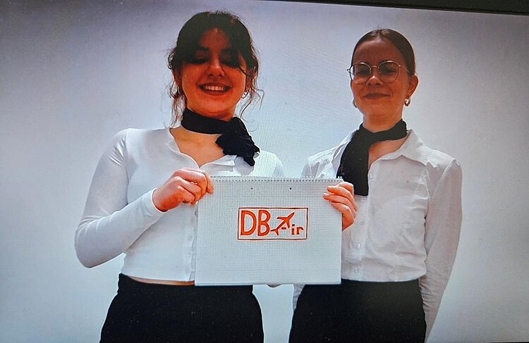Kreative Studierende Zwei als Flugbegleiterinnen verkleidete Auszubildende mit weißem Hemd, Halstuch und dunkler Hose, halten lächelnd ein selbst gestaltetes Logo ihrer fiktiven Firma hoch.