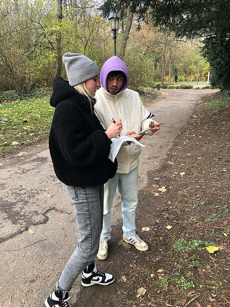 Botanische Exkursion Eine Schülerin und ein Schüler halten einen Tannenzapfen hoch und machen sich, mithilfe der Bestimmungs-App, Notizen auf einem Zettel.