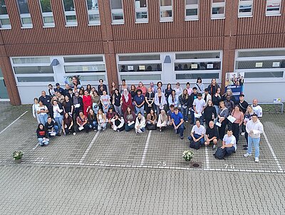 Ein großes Gruppenfoto im Hof vor einem Gebäude. Viele Teilnehmerinnen und Teilnehmer stehen und sitzen zusammen, einige halten Zertifikate in den Händen.