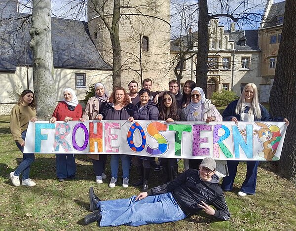 Eine Gruppe von Schülern steht auf einer Wiese vor schlossartigen Gebäuden und hält ein großes Banner mit bunten Buchstaben, auf dem Frohe Ostern steht.