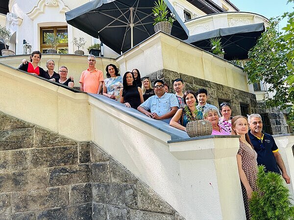 Gruppenfoto von Erwachsenen auf einer Außentreppe vor einem historischen Gebäude mit Terrasse und großen Sonnenschirmen.