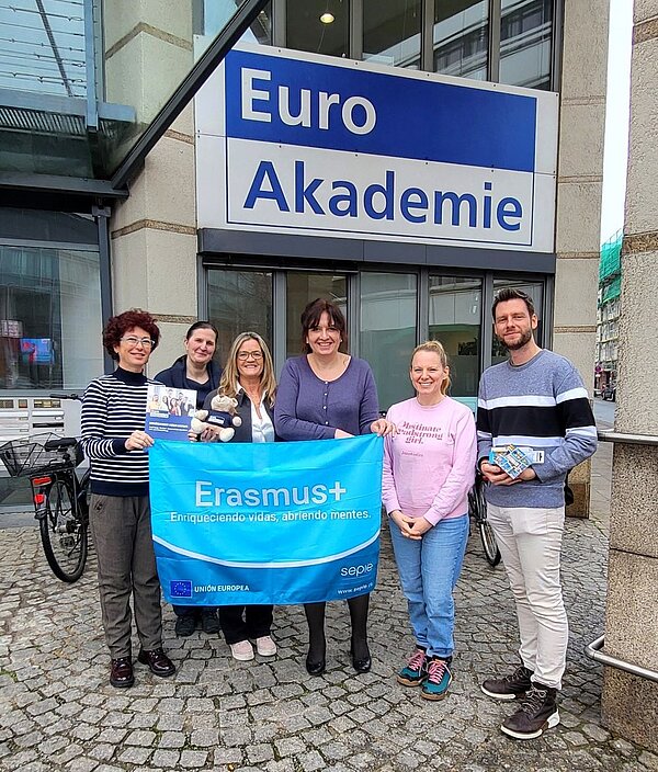 Sechs Personen stehen vor der Euro Akademie und halten eine Erasmus+ Flagge sowie verschiedene Werbematerialien. Die Gruppe besteht aus Männern und Frauen, die lächelnd in die Kamera schauen. Im Hintergrund ist das Schild der Euro Akademie sichtbar.