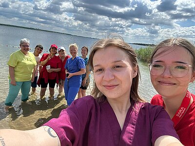 Gruppenfoto am See mit mehreren Personen, die im Wasser stehen. Alle lächeln in die Kamera, es herrscht lockere Stimmung.