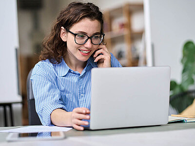 Eine lächelnde Frau mit Brille und blau-weiß gestreiftem Hemd telefoniert, während sie auf einem Laptop arbeitet. Sie sitzt an einem Schreibtisch in einem hellen, modernen Büro.