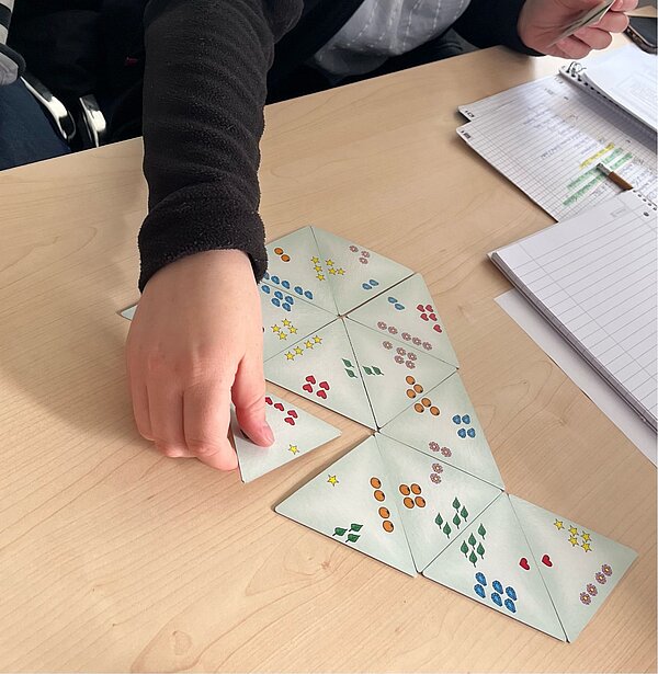 Mathe im Kindergarten? Geht und macht Spaß! Hände bewegen die Spielkarte eines Dominospiels. Das Besondere daran ist, dass die Karte nicht nur zwei, sondern drei Seiten hat, die zu den anliegenden Karten passen müssen.