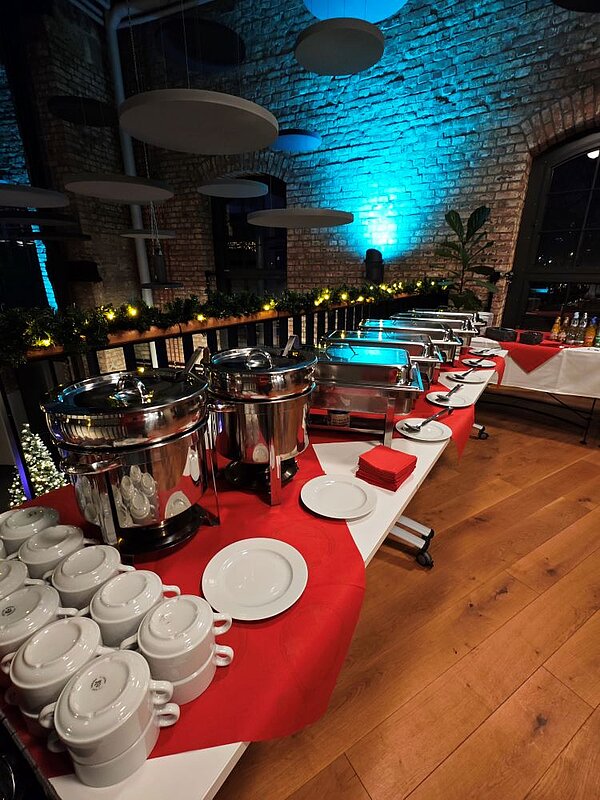 Ein Buffet-Tisch in einem rustikalen Raum mit Ziegelsteinwänden und moderner Beleuchtung. Auf dem Tisch stehen mehrere große, silberne Warmhaltebehälter, daneben Stapel mit weißen Porzellanschüsseln. Der Tisch ist mit einer roten Tischdecke bedeckt. Im Hintergrund sind dekorative Pflanzen und Lichterketten zu sehen.