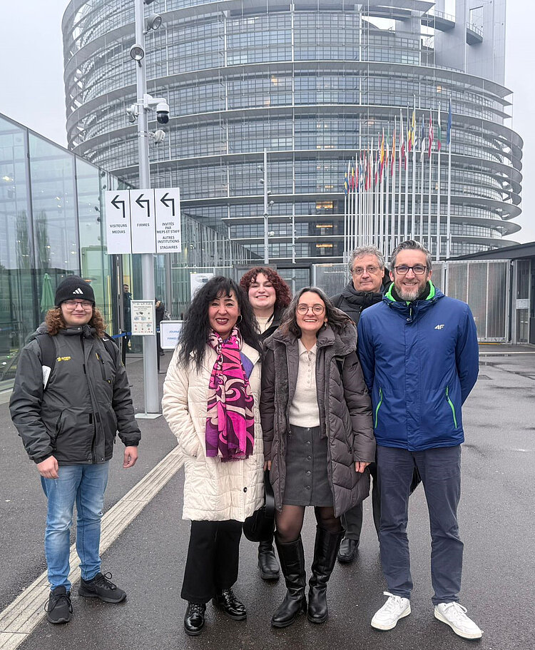 Fünf Personen stehen lächelnd vor dem Eingang des Europäischen Parlaments in Straßburg. Sie sind warm angezogen, was auf kühles Wetter hindeutet. Im Hintergrund sieht man das markante runde Glasgebäude des Parlaments, Flaggenmasten mit verschiedenen Flaggen und ein Hinweisschild mit Richtungsangaben für Besucher, Medien und Abgeordnete. Der Boden ist nass, was auf Regen hindeuten könnte.