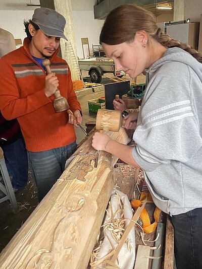 Zwei Personen bearbeiten in einer Werkstatt einen Holzstamm mit Hammer und Meißel. Der Mann links trägt eine graue Kappe und ein orangerotem Pullover, die Frau rechts trägt eine graue Kapuzenjacke. Im Hintergrund sind Werkzeuge, Materialien und ein Autoanhänger zu sehen.