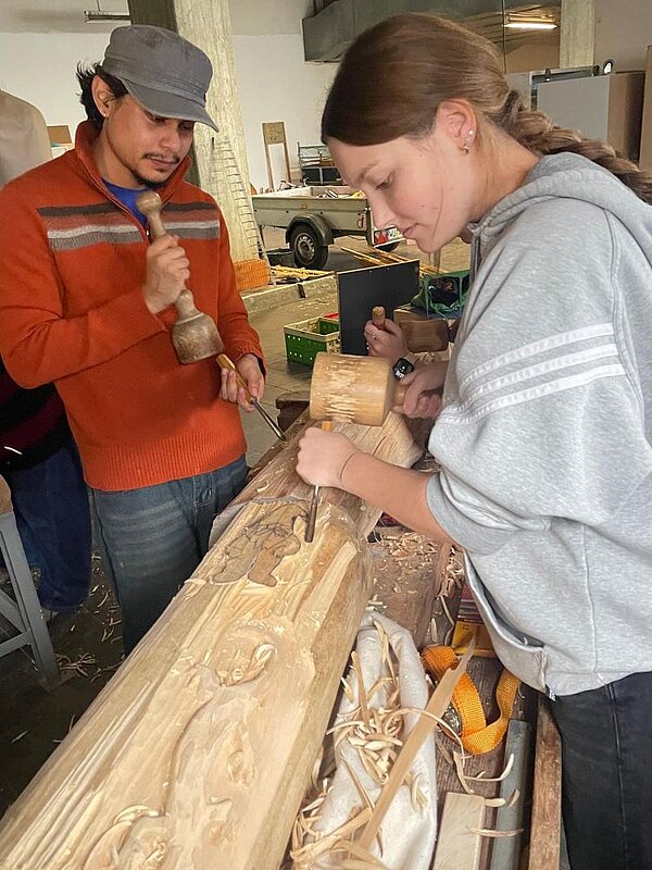 Zwei Personen bearbeiten in einer Werkstatt einen Holzstamm mit Hammer und Meißel. Der Mann links trägt eine graue Kappe und ein orangerotem Pullover, die Frau rechts trägt eine graue Kapuzenjacke. Im Hintergrund sind Werkzeuge, Materialien und ein Autoanhänger zu sehen.