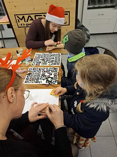 Zwei junge Frauen mit Weihnachtsaccessoires bemalen Kindern die Hände mit Glitzertattoos; auf dem Tisch liegen viele Motiv-Vorlagen, Dosen mit Glitzer und Lack.