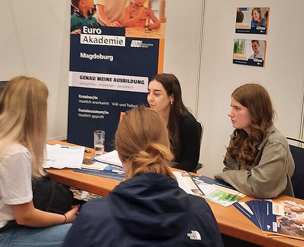 Vocatium Messe Magdeburg 2024 Vor einem Messe-Roll-up sitzen Schülerinnen mit einer jungen Frau an einem Tisch und sind in ein Beratungsgespräch verwickelt. Auf dem Tisch liegen unterschiedliche Infobroschüren.