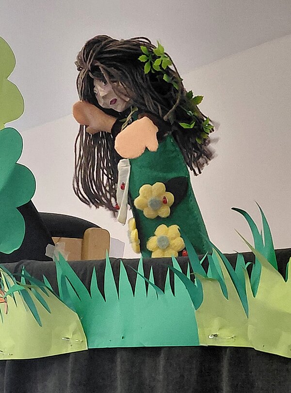 Handpuppe mit langen, dunklen Wollhaaren und grünen Blättern im Haar. Die Figur trägt ein grünes Gewand mit aufgenähten gelben und roten Blumen. Im Vordergrund sind ausgeschnittene Papierformen in Grün zu sehen, die Gras und einen Baum darstellen. Die Szene wirkt wie ein Bühnenbild für ein Puppentheater.