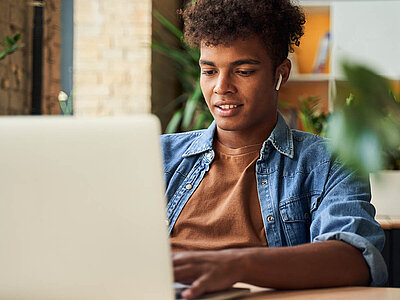 Ein junger Mann in Jeanshemd sitzt vor einem Laptop und hat dabei kabellose, weiße Ohrhörer in den Ohren.