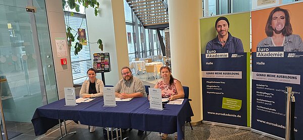 Drei Mitarbeitende sitzen an einem Informationsstand der Euro Akademie Halle; daneben stehen große Roll-ups mit Ausbildungsangeboten.
