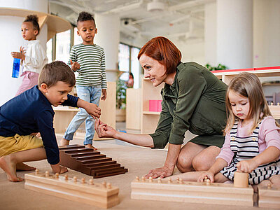 Eine junge Frau spielt in einem großem Raum mit kleinen Kindern auf dem Boden mit verschiedenen Holzspielen. Sie reicht einem kleinen Jungen lächelnd eine Spielfigur, während andere Kinder auch in ihrer Nähe sind. Die Szene spielt sich wahrscheinlich im Umfeld einer Kindertagesstätte ab.