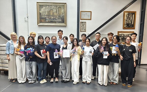 Herzlicher Abschied, freudiger Anfang: Pflege-Absolvent*innen geehrt und neue Auszubildende willkommen geheißen Gruppenfoto von jungen Erwachsenen und einer Lehrkraft, die lächelnd vor einer Wand stehen, an der ein Gemälde und historische Fotos hängen. Die Personen halten Zertifikate und kleine Geschenke in den Händen.