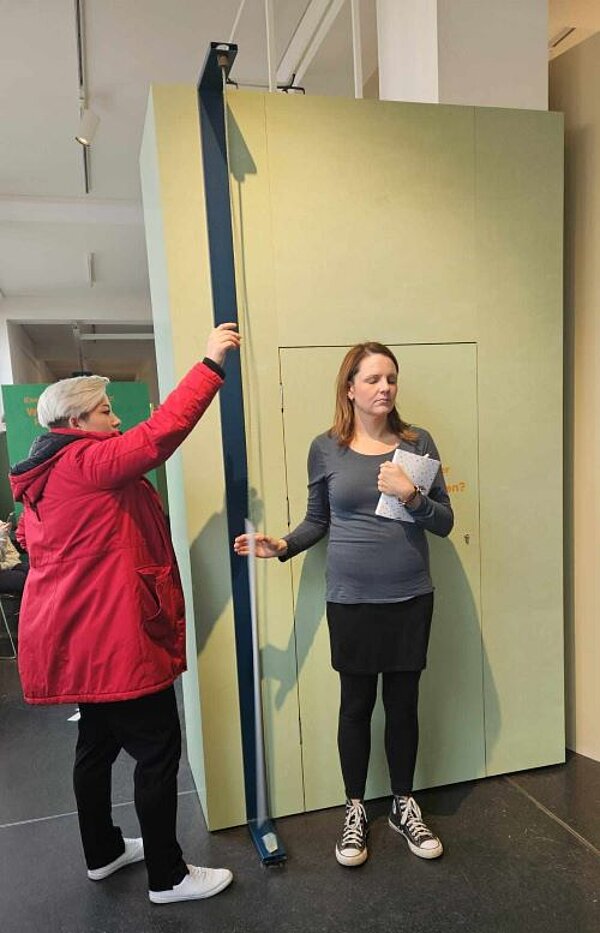 Bewegung und Balance im Hygienemuseum Zwei Frauen an einer Mitmachstation in einem Museum. Eine der beiden Frauen hält eine hohe Leiste fest, an der ein langes Drahtseil angebracht ist. Die andere Frau mit geschlossenen Augen mit dem Rücken zur Wand neben der Leiste und versucht ein an dem Draht hängendes Papier zu greifen.