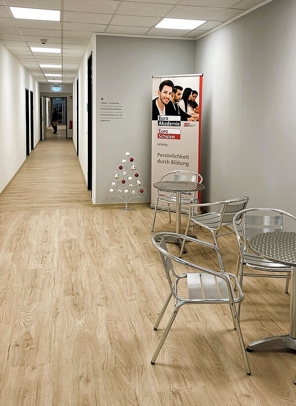 Heller Flur mit hellen Holzboden, Metalltischen und -stühlen, einem Roll-up der „Euro Akademie / Euro Schulen Leipzig – Persönlichkeit durch Bildung“ und einem kleinen weißen Deko-Tannenbaum, Blick in den langen Gang mit Türen.