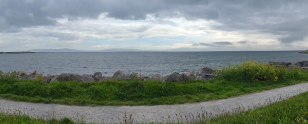 Ein langgezogenes Bild der Küste bei Galway ist zu sehen mit einem Radweg ganz vorne, dahinter Wiese und noch etwas dahinter beginnt das Meer mit einem kleinen Strand.