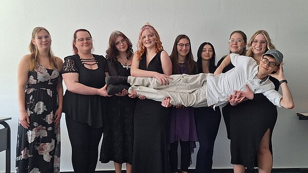 Mottowoche der Abschlussklassen Sozialassistent*in und Erzieher*in Neun junge Frauen in eleganten Kleidern halten lachend einen Mitschüler waagerecht in die Kamera. Alle lächeln – der Junge liegt entspannt in ihren Armen.