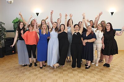 Absolventinnen und Absolventen in feierlicher Kleidung posieren mit jubelnden, nach oben gehobenen Armen für ein Gruppenfoto im großen Saal mit Holzparkett.