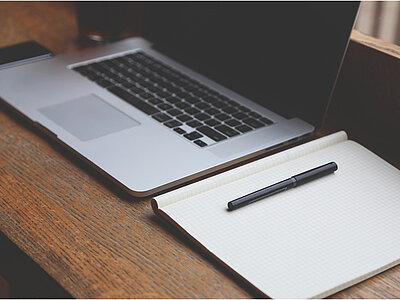 Ein Laptop und ein Notizbuch liegen auf einem Holztisch. Der Laptop ist geöffnet und zeigt die Tastatur und das Trackpad. Daneben liegt ein schwarzer Stift auf dem Notizbuch, das ebenfalls geöffnet ist und leere Seiten zeigt.