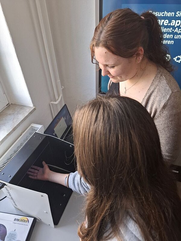 Eine Person mit langem Haar hält eine Hand in ein Gerät, das auf einem Tisch steht. Sie scheint eine Untersuchung oder Messung durchzuführen, während eine andere Person sie anleitet.