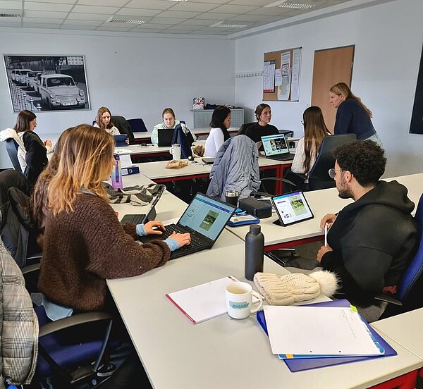 Ein Gruppenarbeitsraum mit mehreren Personen, die an Tischen sitzen und an Laptops oder Tablets arbeiten. Die Atmosphäre wirkt konzentriert und kollaborativ. Im Hintergrund ist eine Person zu sehen, die einer anderen Person etwas erklärt oder zeigt. Der Raum ist modern eingerichtet, mit einem großen Bild an der Wand und einem Whiteboard.