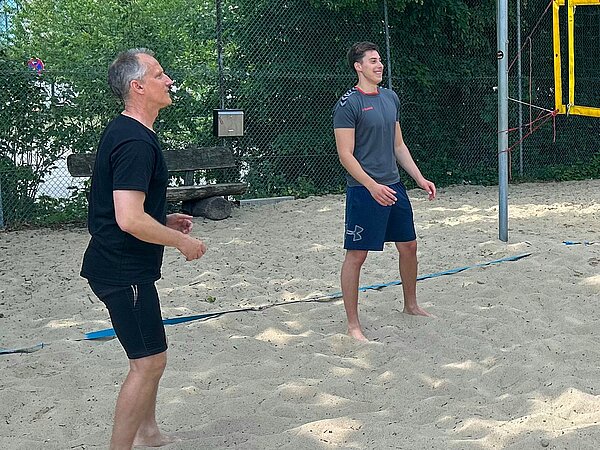Beachvolleyball mitten in Hannover Ein Lehrer und ein Schüler der Euro Akademie Hannover befinden sich mitten im Beachvolleyballspiel. Beide stehen barfuß im Sand, der Schüler lacht, während der Lehrer gespannt nach oben blickt.