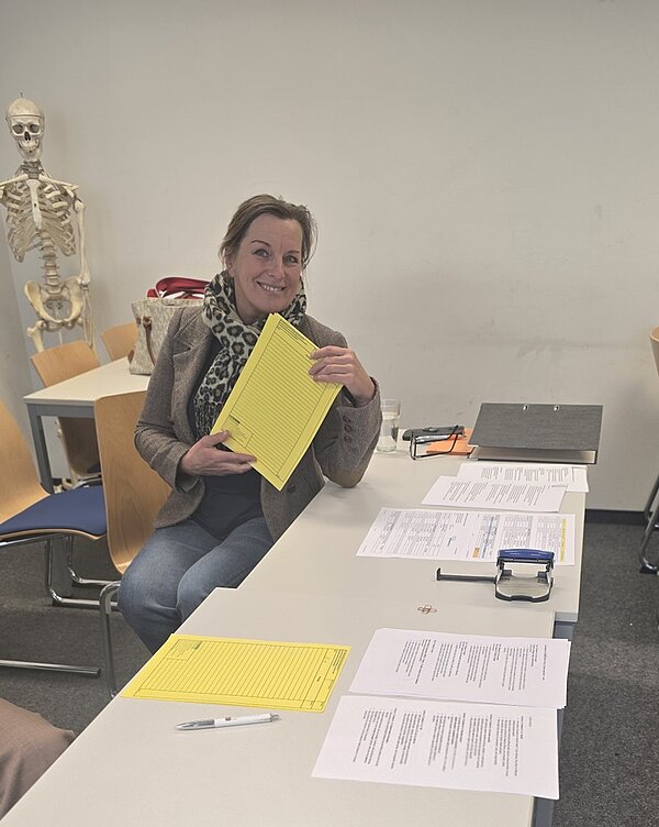 Eine Mitarbeiterin mit kurzem, hellbraunem Haar sitzt an einem Tisch in einem Raum mit weißen Wänden. Sie trägt einen hellen Mantel und einen gemusterten Schal. In der Hand hält sie ein gelbes Formular, das einen Großteil ihres Gesichts verdeckt. Auf dem Tisch liegen mehrere weiße Blätter mit Text, ein Stempelgerät, ein schwarzer Aktenordner und ein Kugelschreiber. Im Hintergrund steht ein Skelettmodell neben mehreren leeren Holzstühlen.