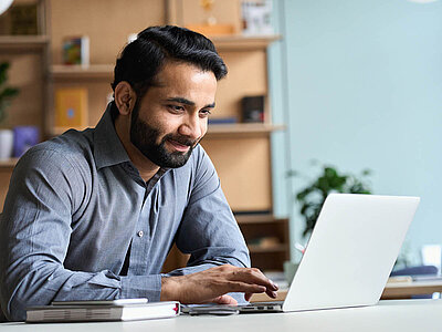 Ein lächelnder Mann mit Bart und einem grauen Hemd sitzt an einem Schreibtisch und arbeitet konzentriert an einem Laptop. Auf dem Tisch liegen ein Notizbuch und ein Stift. Der Mann wirkt entspannt und zufrieden. Im Hintergrund sind Regale mit Büchern und Ordnern sowie eine Zimmerpflanze zu sehen, was auf ein Büro oder Homeoffice hindeutet. Der Raum wirkt freundlich und gut beleuchtet.