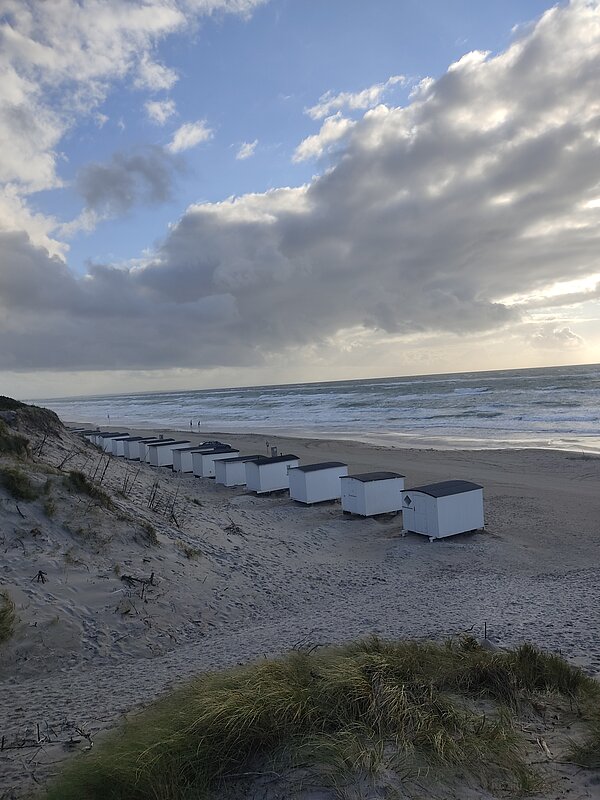Erfahrungsbericht Erasmus – Hygge von Janice Weßel Eine Küstenlandschaft mit einer Reihe kleiner, weißer Badehäuschen auf einer Sanddüne. Die raue Nordsee erstreckt sich im Hintergrund, während große Wolken am Himmel vorbeiziehen. Dünengras bedeckt teilweise den Vordergrund.