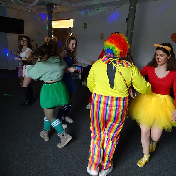Eine Party mit vielen kostümierten Personen, darunter ein Clown mit einer bunten Perücke, eine Frau im roten Oberteil und gelbem Tutu sowie weitere Gäste in farbenfrohen Outfits. Die Atmosphäre ist ausgelassen mit tanzenden und feiernden Menschen.