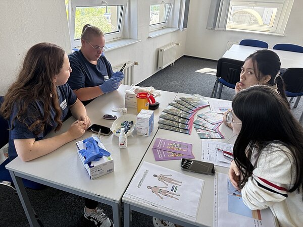 Tag der offenen Tür an der Euro Akademie Bitterfeld-Wolfen Zwei weibliche Auszubildende tragen Euro Akademie-T-Shirts in marineblau, sitzen an einem großen Tisch, auf dem allerlei Hygiene- und medizinische Artikel liegen, ihnen gegenüber sitzen zwei Besucherinnen und schauen ihnen zu.