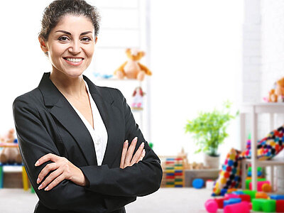 Auf dem Bild ist eine Frau im Anzug zu sehen. Im Hintergrund befinden sich bunte Spielzeuge, darunter Bausteine, Stofftiere und Pflanzen. Die Szenerie wirkt lebhaft und farbenfroh.