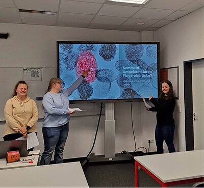 Drei Schülerinnen präsentieren gemeinsam eine Folie mit großen Fingerabdrücken. Eine von ihnen zeigt auf einen roten Fingerabdruck. Der Titel der Präsentation lautet: „Sammeln Ampelarmaturen Fingerabdrücke?“.