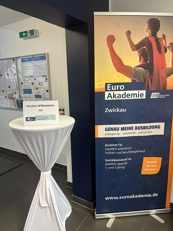 In einem Flur steht neben einem Roll-up-Banner der Euro Akademie ein weißer Stehtisch mit einem Herzlich-Willkommen-Schild.