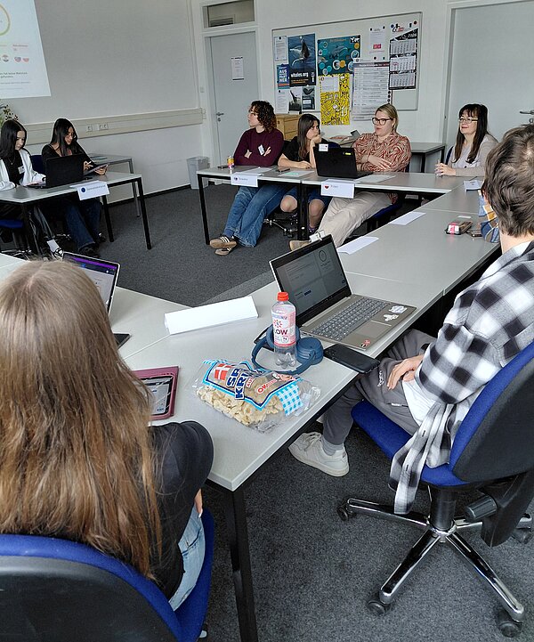 Mehrere Personen sitzen in einem Klassenzimmer, in dem die Tische zu einem Viereck angeordnet sind; auf den Tischen stehen Laptops und Namensschilder.